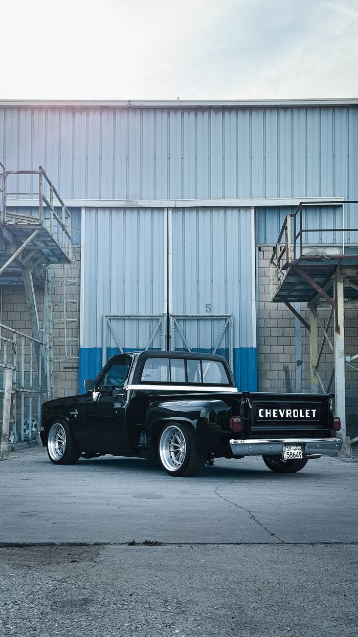 Back view of a vintage black Chevrolet pickup truck parked in an urban industrial area.