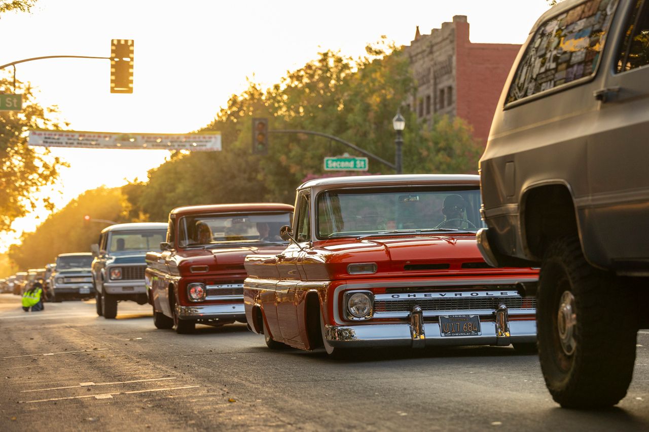 gallery-1 Vintage Chevrolet trucks cruising down a sunlit city street during a retro car show.