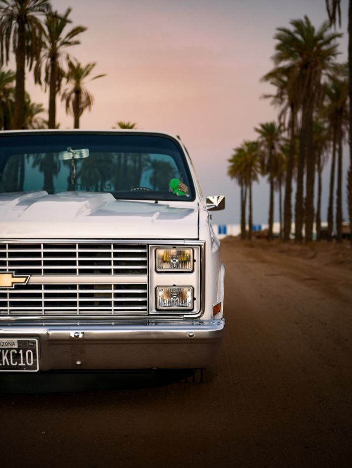 gallery-2 Vintage Chevrolet truck driving on a palm-lined desert road at sunset.