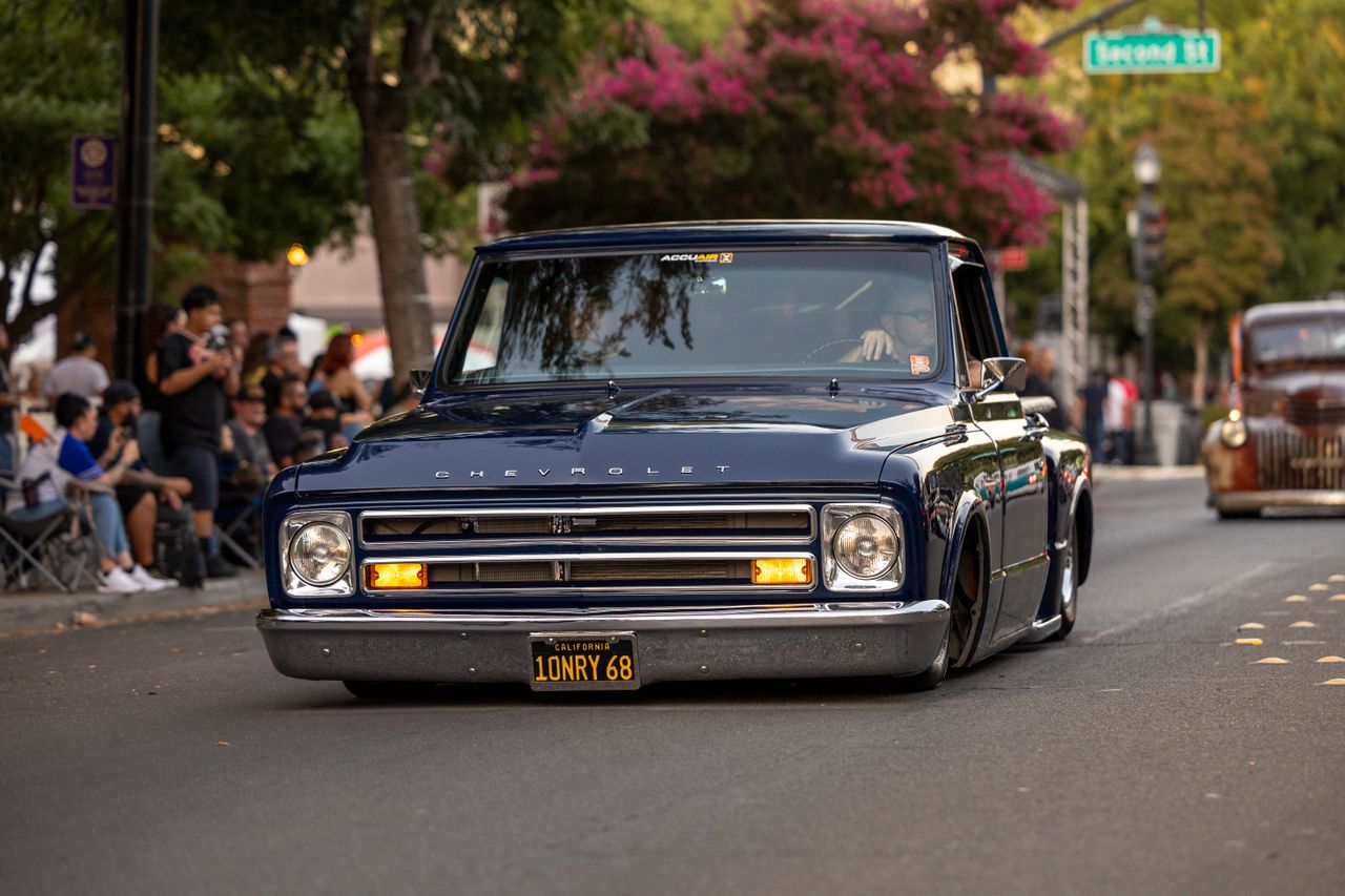 hero-img-01 Blue Chevrolet truck cruising down street during car show with audience under trees.