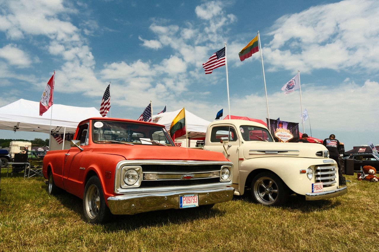 gallery-6 A colorful outdoor exhibit of vintage Chevrolet and Ford trucks amidst flags and tents at a car show.